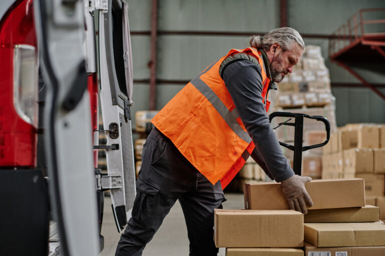 Mature worker loading cardboard containers in truck during his work in warehouse